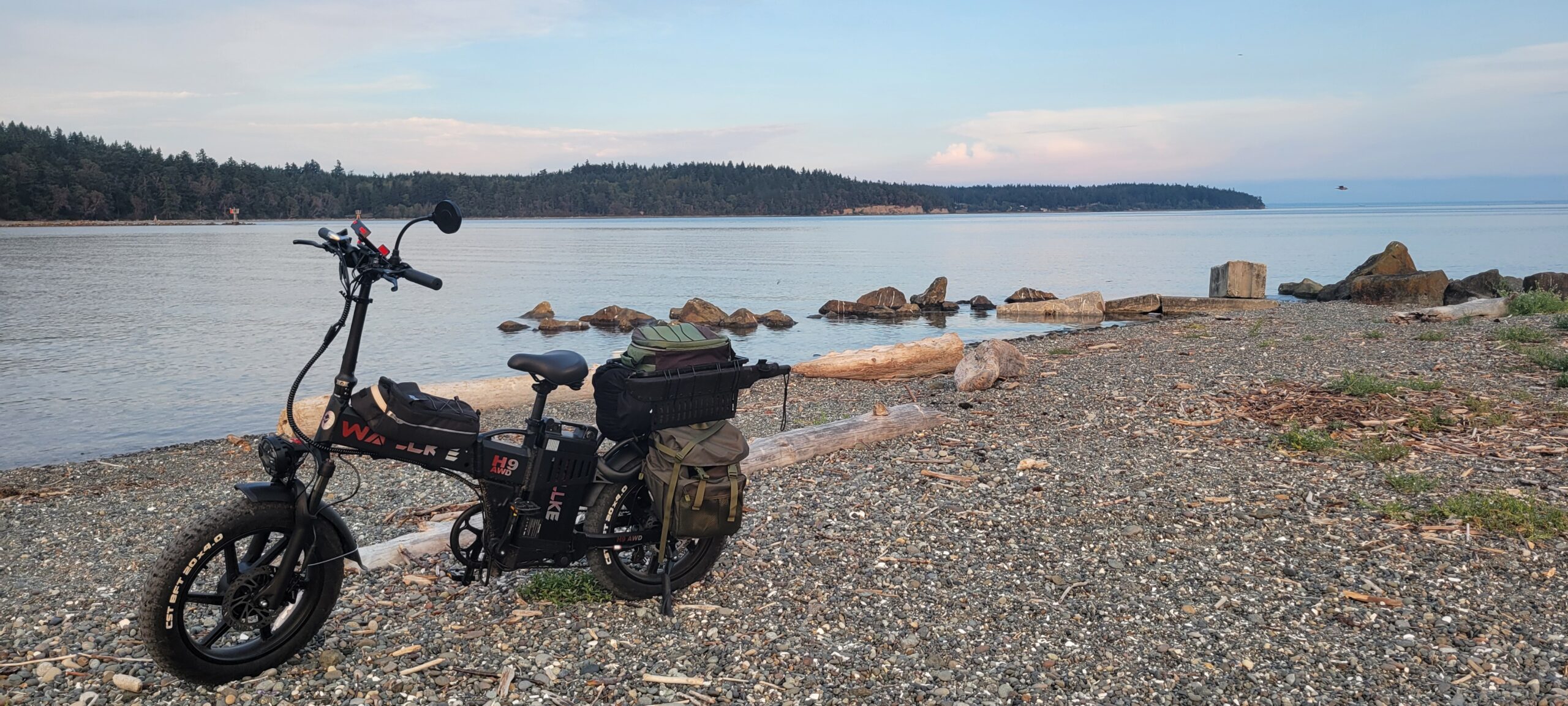 Wallke H9 AWD electric fat-tire bike resting on a rocky beach with the ocean and distant shoreline in the background during sunset